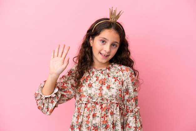 Pequena princesa caucasiana com coroa isolada no fundo rosa saudando com a mão com expressão feliz