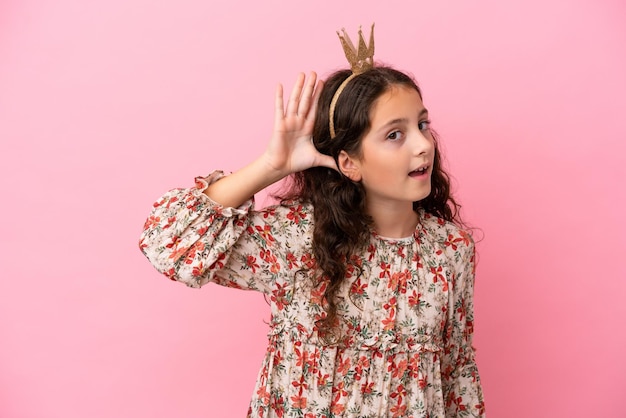 Pequeña princesa caucásica con corona aislada de fondo rosa escuchando algo poniendo la mano en la oreja