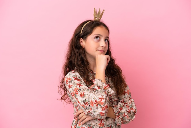 Pequeña princesa caucásica con corona aislada en un fondo rosa que tiene dudas y piensa