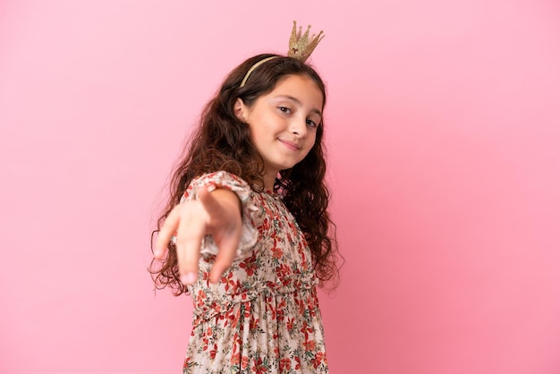 Pequeña princesa caucásica con corona aislada sobre fondo rosa apuntando al frente con expresión feliz