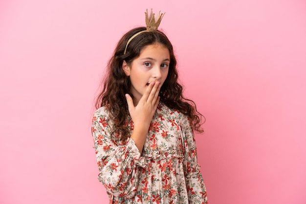 Pequeña princesa caucásica con corona aislada sobre fondo rosa feliz y sonriente cubriendo la boca con la mano