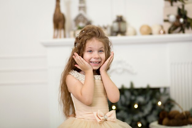 Pequeña princesa linda niña en un elegante vestido ríe y posa cerca del árbol de Navidad