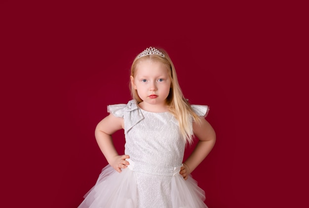 Pequeña princesa rubia hermosa que baila en el vestido blanco y de plata de lujo aislado en fondo rojo. Cara divertida, diferentes emociones