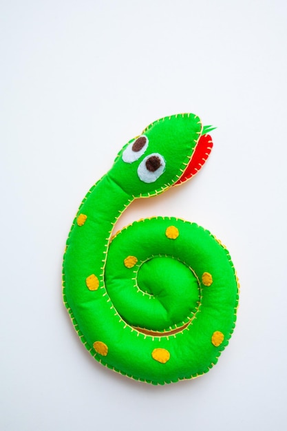 Una pequeña serpiente verde esponjosa con grandes ojos adorables está envuelta alrededor de la mano de una persona Su textura suave y aspecto lúdico la hacen parecer un juguete de peluche acogedor El fondo muestra un cálido