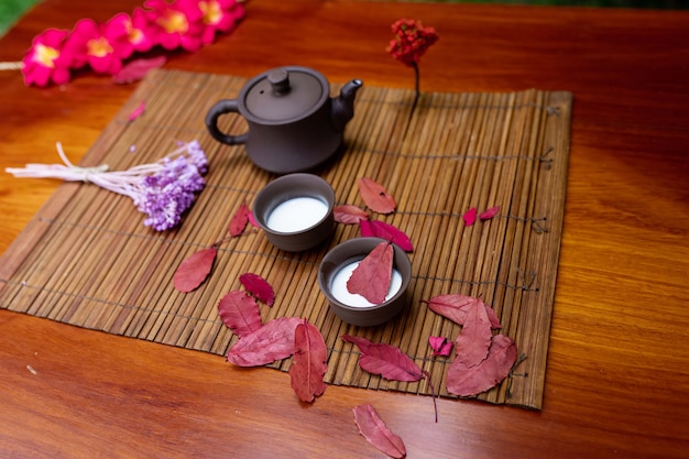 Una pequeña tetera de arcilla con dos tazas para bebidas de pie sobre una estera entre sábanas rojas con una ramita de madera de lavanda y flores de magnolia roja