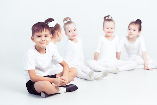 Pequeñas bailarinas y kid ballerun haciendo ejercicios y sentados en el suelo en la clase de ballet blanco.