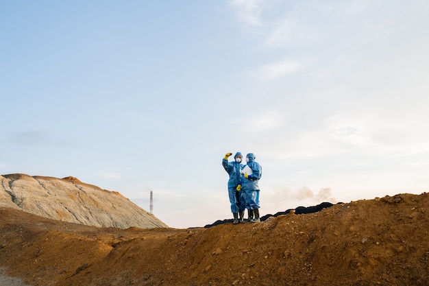 Pequeñas figuras de dos ecologistas de pie en la cima de una colina durante la investigación científica de un territorio abandonado con suelo y aire contaminados