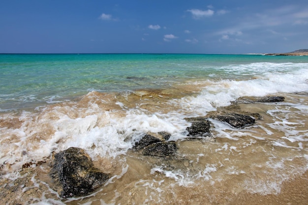 Pequenas ondas do mar esmagando rochas negras na praia, belas águas cristalinas azuis no fundo. Karpass, Chipre do Norte