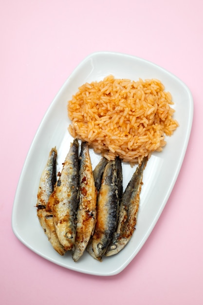 Pequeñas sardinas fritas con arroz con tomate en un plato blanco sobre mesa rosa