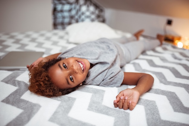 Pequeño afroamericano que miente en una cama, mirando la cámara.