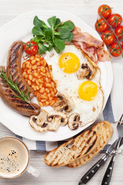 Pequeno-almoço inglês: ovos fritos, salsichas, bacon, feijão, torradas, tomates e xícara de café em uma mesa de madeira.