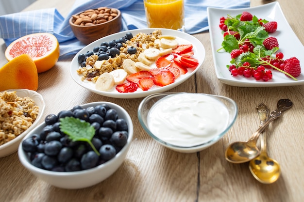 Pequeno-almoço saudável servido com prato de iogurte muesli mirtilos, morangos e banana. Mesa de manhã granola amêndoas bagas suco de frutas cítricas e ervas verdes.