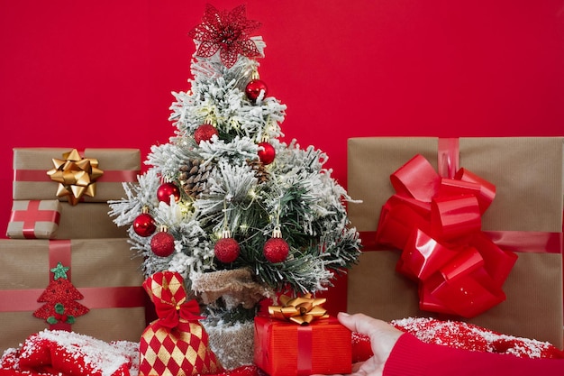 Pequeño árbol de Navidad decorado y mano femenina que ofrece un presente Copia espacio sobre fondo rojo