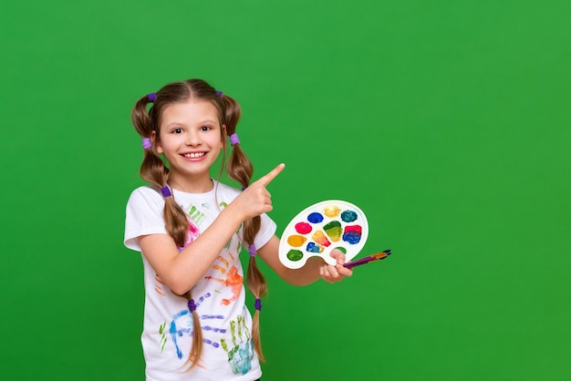 Un pequeño artista con una paleta de colores apunta a su anuncio sobre un fondo verde aislado Cursos de arte para niños