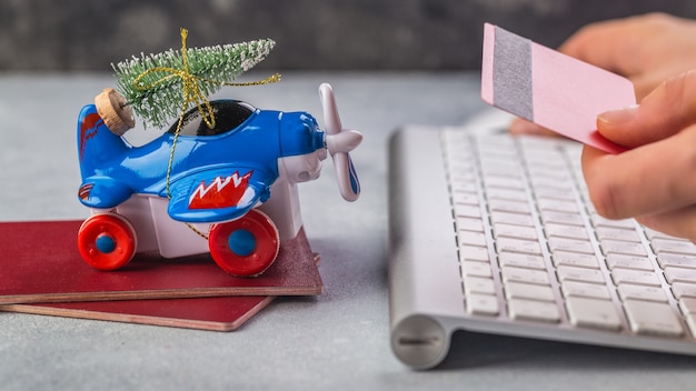 Pequeño avión con árbol de Navidad, pasaportes, teclado en mano gris mujer está tomando tarjeta de crédito