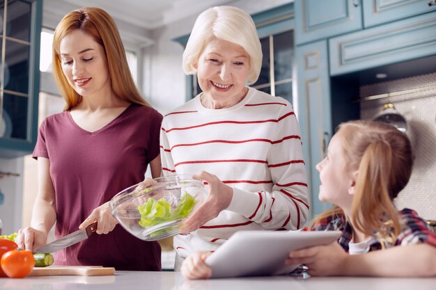 Pequeño ayudante. Encantadora niña sentada en la encimera de la cocina y mostrando una tableta con una receta a su madre y abuela mientras las mujeres hacen ensalada