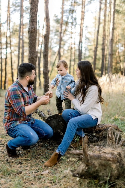 Pequeño bebé adorable lindo, divirtiéndose mientras se balancea en el árbol de registro, tomados de la mano de su madre bastante atractiva y su padre barbudo guapo en el bosque de otoño al aire libre