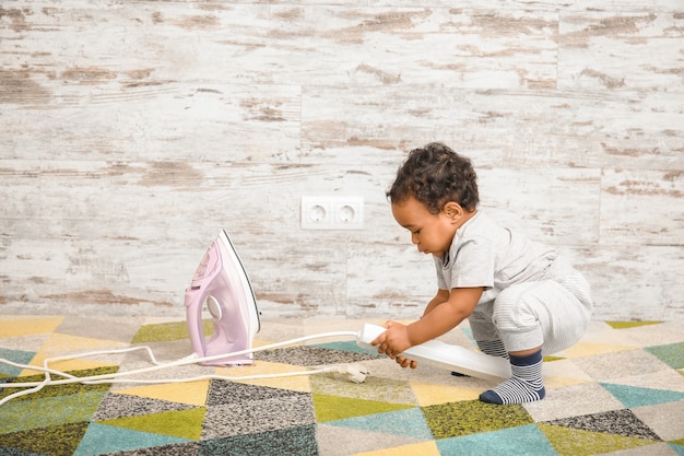 Pequeño bebé afroamericano jugando con plancha en casa. Niño en peligro