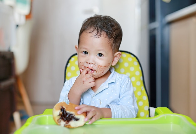 El pequeño bebé asiático feliz que se sienta en niños preside el pan interior de la consumición con el chocolate relleno