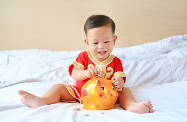 Pequeño bebé asiático sonriente que pone algunas monedas en una hucha en cama.