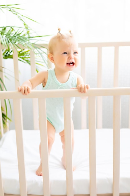 Pequeño bebé divertido parado en una cuna en una guardería blanca con una ventana Una niña pequeña con un traje de algodón El bebé es travieso no quiere dormir en su cuna El problema del sueño de los niños