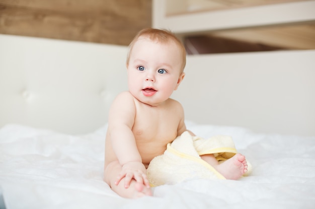 Pequeño bebé divertido que ríe cuando se sienta en la cama con la toalla