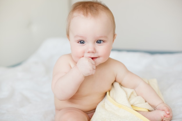 Pequeño bebé divertido que ríe cuando se sienta en la cama con la toalla