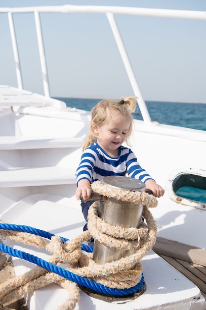 Pequeño bebé o niño lindo con cara feliz y cabello rubio en camisa despojada jugando con cuerda en barco blanco verano soleado al aire libre sobre fondo natural