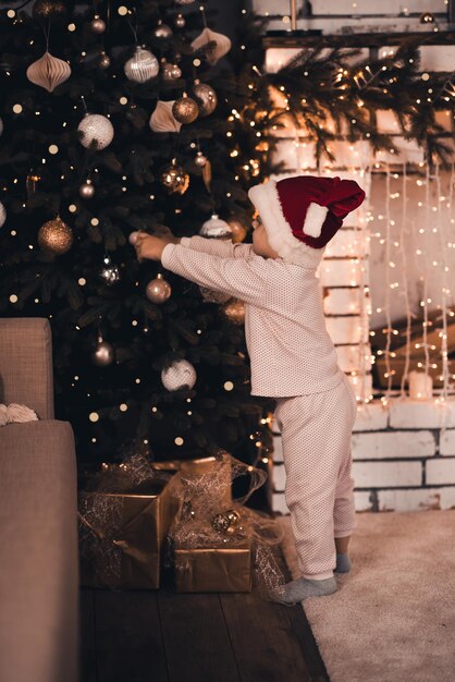 Pequeño bebé en pijama y gorro de santa claus decoran el árbol de Navidad