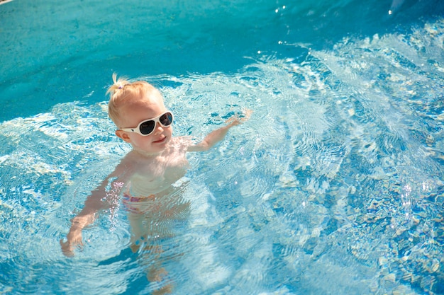 Pequeño bebé rubio lindo con las gafas de sol que salpican feliz en la piscina con agua azul clara.