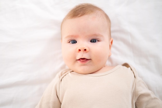 Un pequeño bebé sano con un traje de algodón yace boca arriba en una cama con ropa de cama blanca en el dormitorio de su casa mira a la cámara sonríe un gran retrato del niño