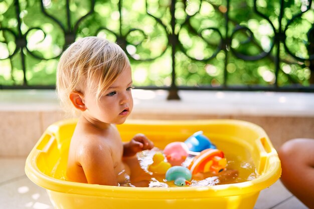 El pequeño bebé se sienta en una palangana con agua y juguetes en el balcón