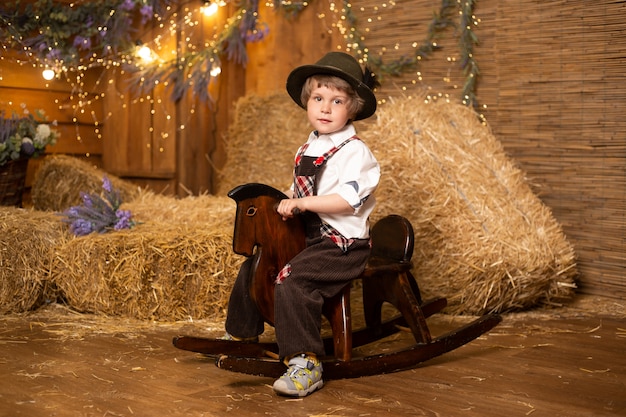 Pequeño bebé sonriente que se sienta en el caballo del juguete que lleva el traje retro en granja con las gavillas de paja