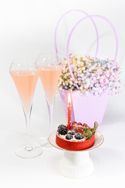Pequeno bolo de celebração com vela acesa dois copos de vinho espumante e um cesto com flores
