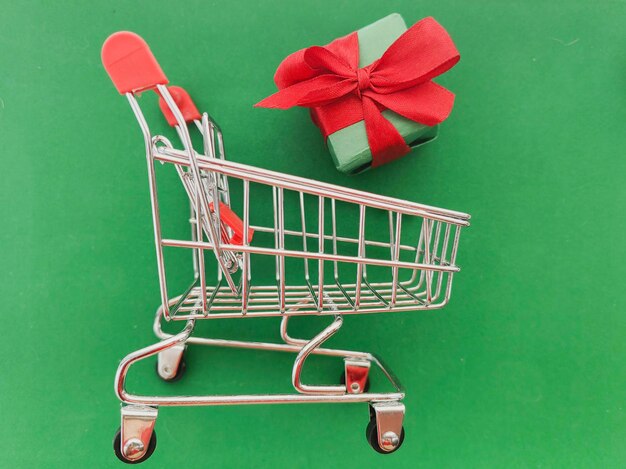 Un pequeño carrito de compras con un lazo rojo se sienta al lado de una caja verde con un fondo verde