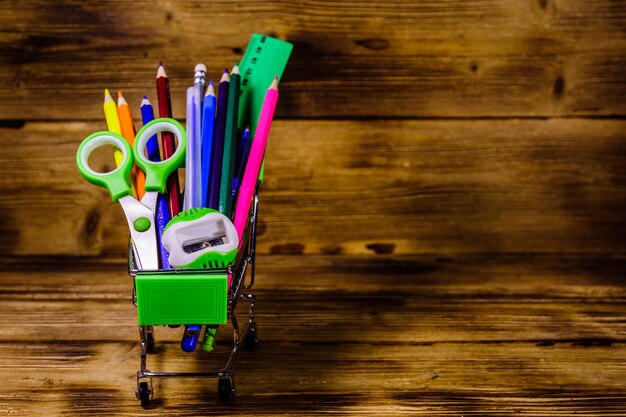 Pequeño carrito de compras con papelería escolar diferente sobre fondo de madera rústica Concepto de regreso a la escuela