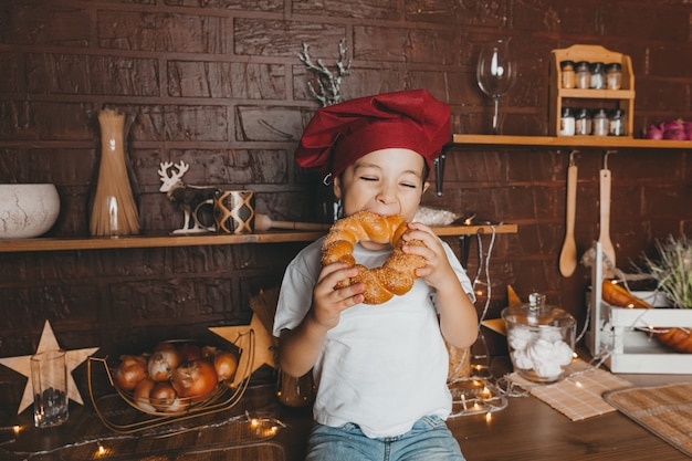 Pequeño chef. Niño con gorro de cocinero con pasteles, bollos, pan y bagels. Niño comiendo un bagel.