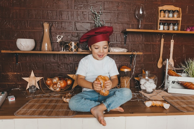 Pequeño chef. Niño con gorro de cocinero con pasteles, bollos, pan y bagels. Niño comiendo un bagel.