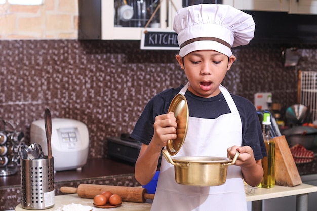 El pequeño chef sorprendido abre la cacerola o la olla coreana contra el fondo de la cocina