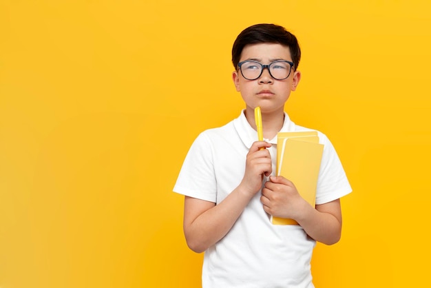 un pequeño colegial asiático sosteniendo un bolígrafo y libros y pensando sobre un niño coreano pensativo de fondo amarillo