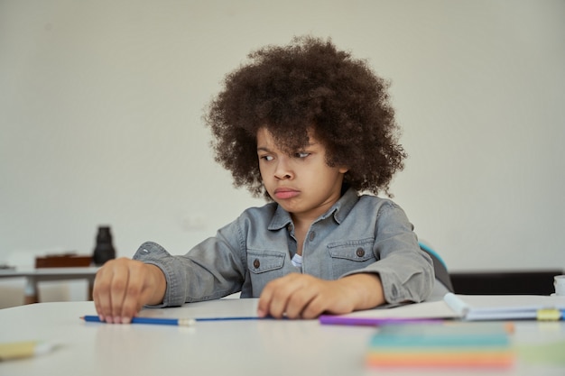 Pequeño colegial descontento con cabello afro que parece triste mientras está sentado en la mesa en la escuela primaria