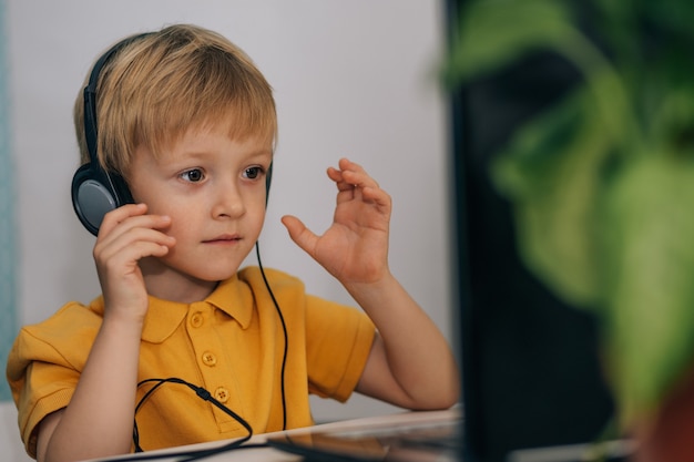 Un pequeño colegial interesado con auriculares está sentado en la mesa de la casa y mira un video tut