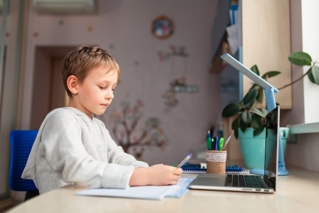 Pequeño colegial lindo que estudia en casa haciendo los deberes escolares. Libros de formación y cuaderno sobre la mesa. Educación a distancia en línea