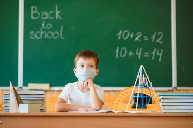 Pequeño colegial lindo sentado en el aula con una máscara