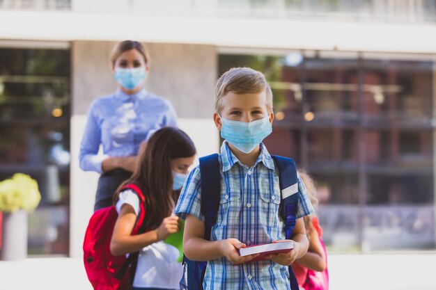 El pequeño colegial con una máscara protectora se para frente a la escuela El niño con una mochila sostiene un libro