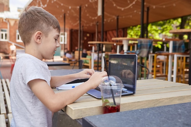 El pequeño colegial se sienta al aire libre, escribe en el teclado de la computadora portátil. niño caucásico sonriente sentado en la mesa en la veranda del café, estudiando con la computadora, haciendo la tarea. aprendizaje a distancia, educación en línea
