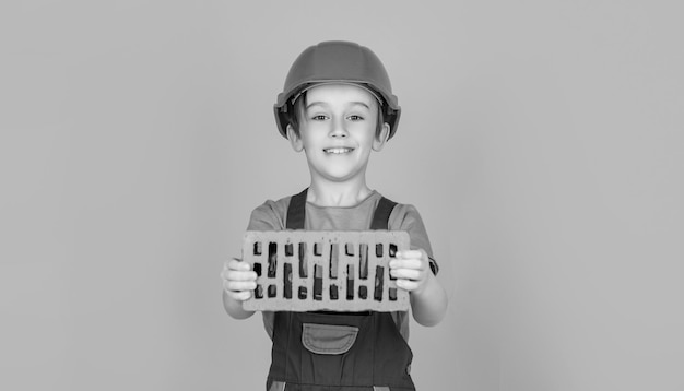 Pequeno construtor de capacete criança vestida como um construtor de operário menino em um capacete de construção tem um tijolo nas mãos em fundo amarelo criança capacete de construção capacete preto e branco