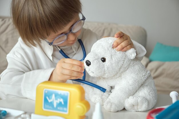 Pequeño doctor en abrigo médico con estetoscopio juega tratamiento ocular para juguete de oso de peluche