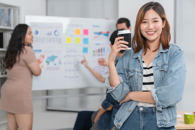 Pequeño emprendedor asiático inteligente de pie retrato confiado con amigos socios reunión informal lluvia de ideas con pizarra blanca y análisis de gráficos de papel de finanzas comerciales en el fondo de la oficina