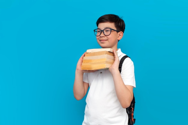 Pequeno estudante asiático de óculos com mochila segurando livros e sorrindo em fundo azul